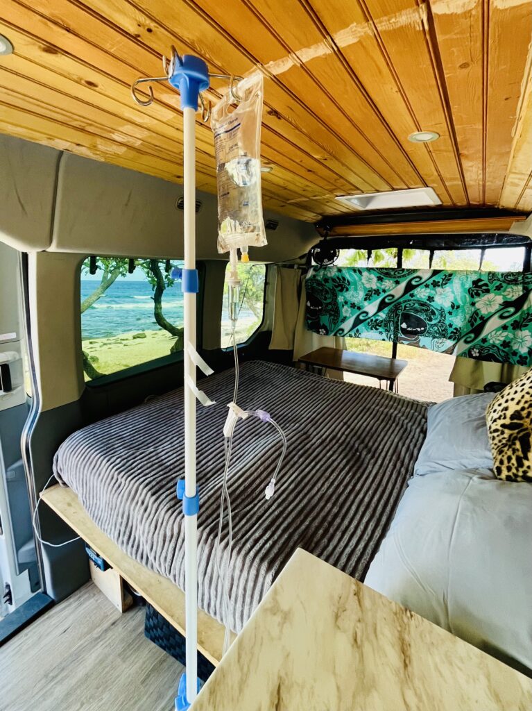 Mobile IV drip setup beside a bed in a camper van parked on the beach in Kailua-Kona, Hawaii, with ocean view through the window